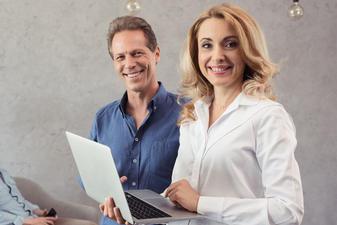 Smiling man and woman holding a laptop in a casual setting.